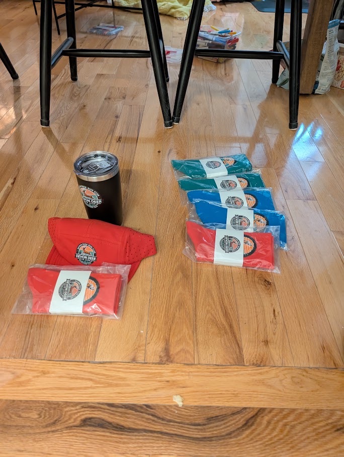 Des chaussettes de course Devon Deer rouges, bleues et vertes sont disposées avec un gobelet isotherme noir et une casquette rouge.