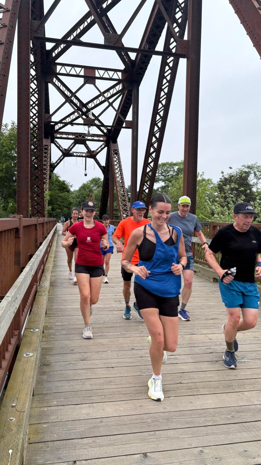A smiling pack of Devon Deer runners moving toward the camera across Fredericton’s railway bridge.