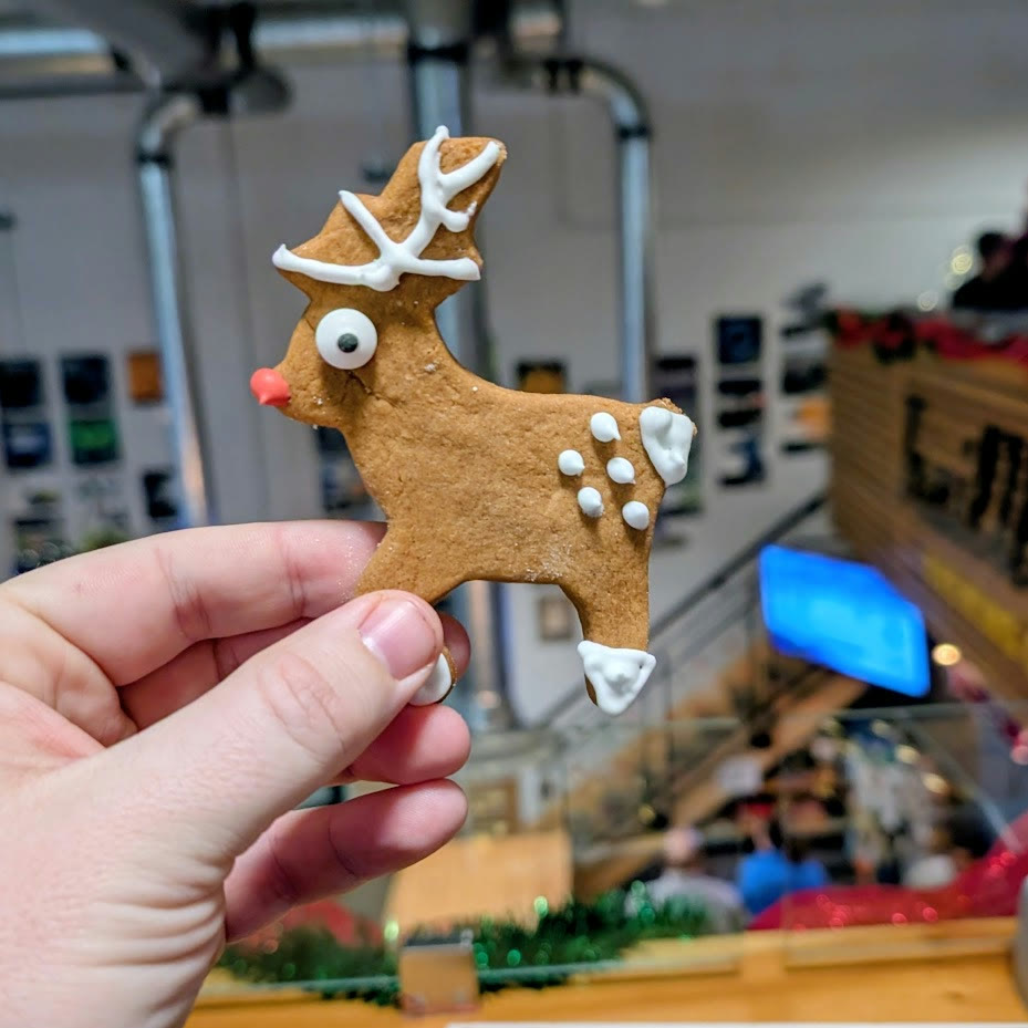 A hand holds a deer-shaped gingerbread cookie decorated with white icing and a red nose at a Devon Deer holiday gathering.